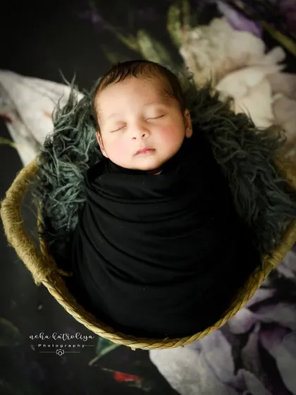 A classic portrait of a baby in a black wrap, sleeping peacefully in a basket. The dark tones create a dramatic and timeless feel.