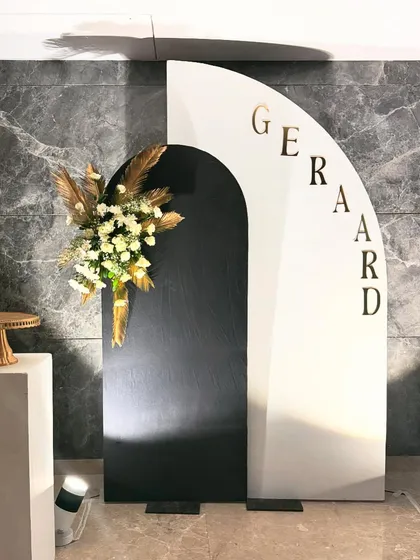 A chic and minimalist birthday backdrop with a modern black and white arched panel. The decor is accented with a spray of dried pampas grass and white flowers, and personalized with the name "Gerard" in gold letters.