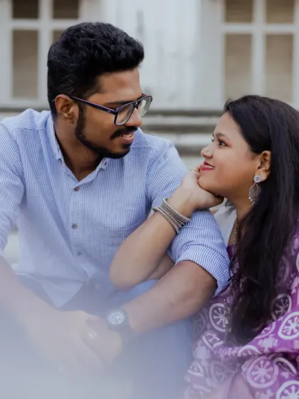 A sweet, candid moment during a pre-wedding shoot at a location with classic architecture. The bride-to-be gently touches her partner's face.