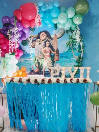 A tropical Moana-themed 10th birthday party. The decor includes a backdrop of the ocean, a colorful balloon garland, and a fringed table skirt to create an island vibe.