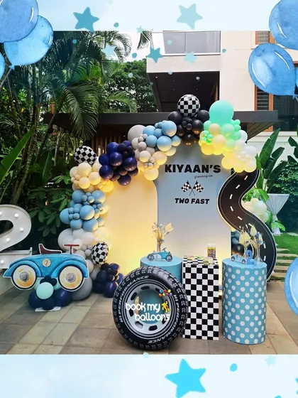 For Kiyaan's "Two Fast" second birthday, we designed a sleek and fun race car theme. The decor features a custom road backdrop, a balloon arch in shades of blue and checkered patterns, and props like a giant tire and a vintage car cutout.