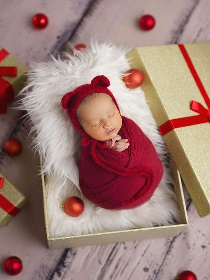 The perfect little gift. This newborn is styled in a festive red wrap and hat, nestled inside a golden gift box for a memorable holiday-themed announcement.