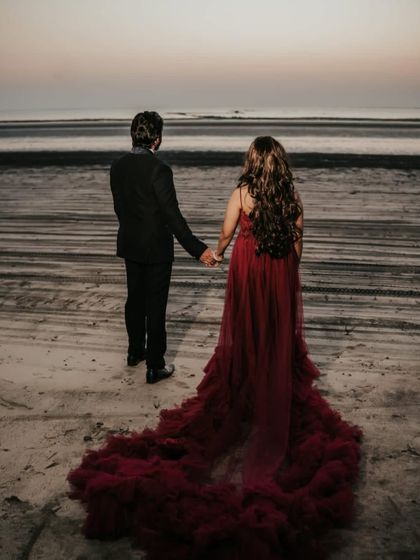 Another angle of the beach photoshoot, showing the full length of the dramatic red trail gown as the couple walks along the shore.