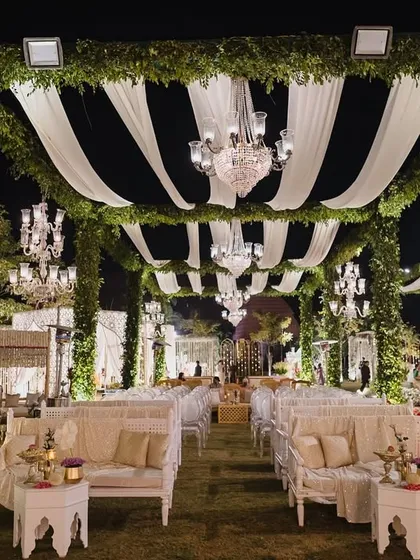 A symphony of florals and light. This elegant outdoor wedding seating is arranged under a canopy of green garlands and white drapes, with crystal chandeliers adding a touch of romance.