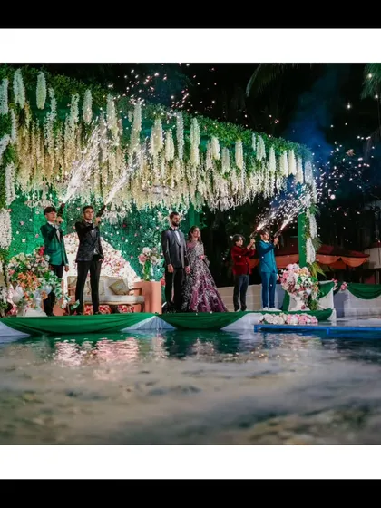 An unforgettable reception moment. The couple stands on a stage set over a pool, complete with a floral backdrop and champagne spray, showcasing a truly spectacular celebration.