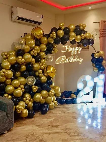 Celebrating a 21st birthday in style. This setup features a dramatic black and gold organic balloon arch, a custom 'Happy Birthday' neon sign, and large, illuminated marquee numbers to mark the special occasion in any home.