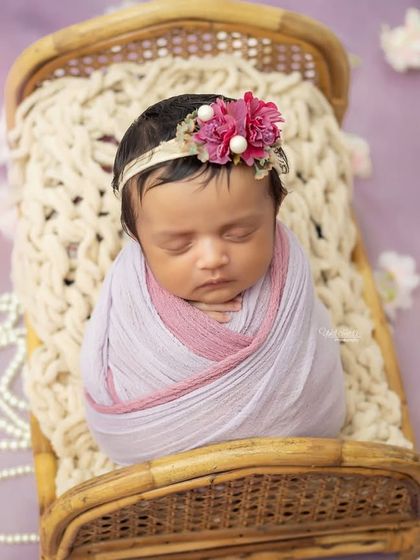 A beautiful newborn girl swaddled in shades of lavender and pink, resting on a tiny bed. The floral headband adds a delicate touch.