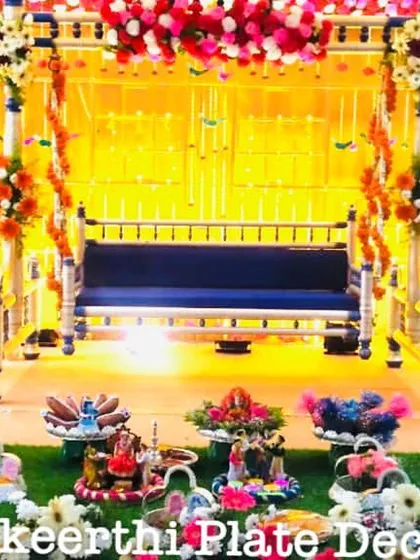 A vibrant baby shower setup at Brookefields, Coimbatore, with a beautiful swing as the centerpiece. The 15 platters on the grass floor are filled with colorful sweets, fruits, and thematic dolls.