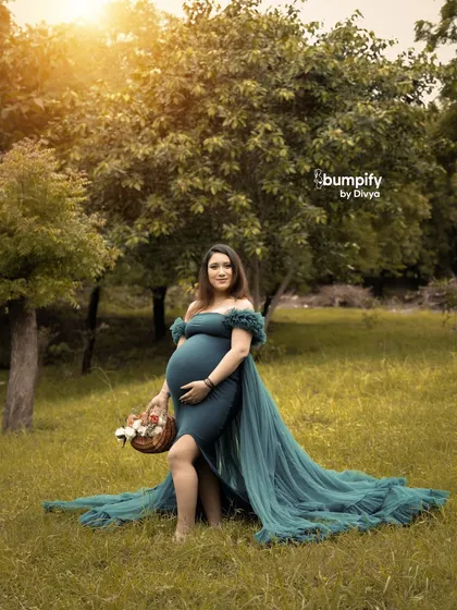 A solo portrait of a radiant mom-to-be in a flowing teal gown, standing in a beautiful green meadow. She holds a basket of flowers, adding a lovely, natural touch to this outdoor maternity photo.