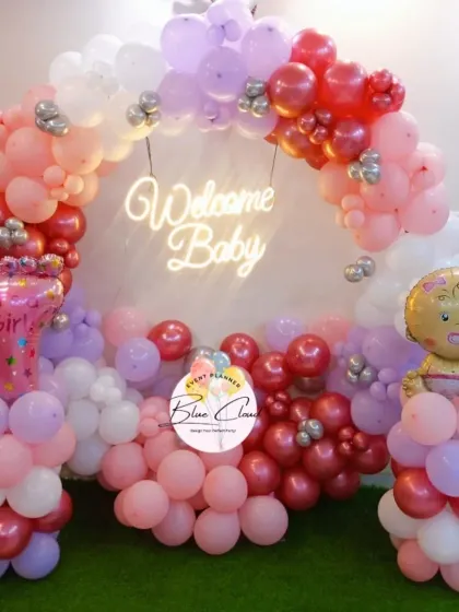 A 'Welcome Baby' girl balloon arch in shades of pink, red, and purple, with a neon sign.