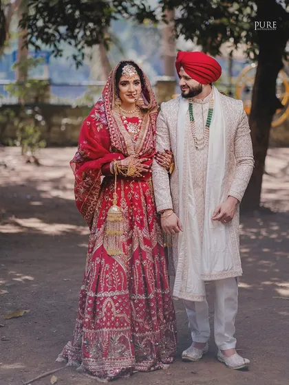 A classic portrait of the bride and groom. The full-length shot showcases their stunning red and white wedding outfits in a natural, outdoor setting.