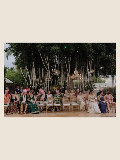 A wide shot of guests seated for an outdoor ceremony under a large tree adorned with hanging chandeliers, capturing the event's elegant and natural ambiance.