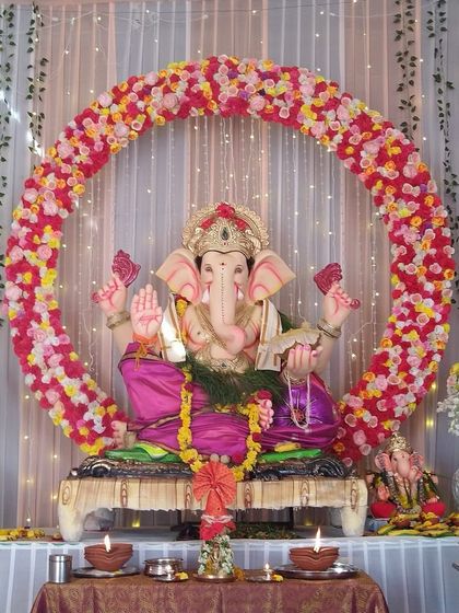 A full circular arch made of pink, yellow, and white flowers creates a cheerful frame for this large Ganpati idol. The simple white curtain with fairy lights provides a soft, glowing background.