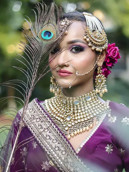 A unique bridal portrait using a peacock feather as a prop. The makeup, with its purple and pink tones, beautifully complements the colors of the feather and the outfit.
