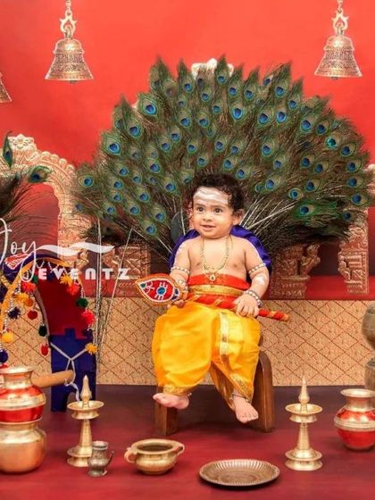 A grand and divine Little Muruga themed photoshoot for kids. This elaborate setup features a peacock feather throne, traditional brass lamps, and temple bells, creating a powerful and culturally rich portrait.