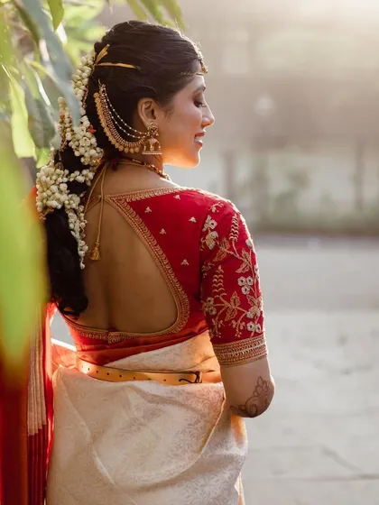 A bride wearing a classic off-white and red Kanchipuram saree. Her custom red blouse features delicate floral embroidery on the back and sleeves.