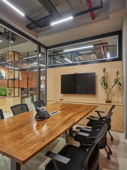 A meeting room with an exposed brick wall and a large wooden table, viewed from the outside. The glass partitions allow for visual continuity with the adjacent reception area.
