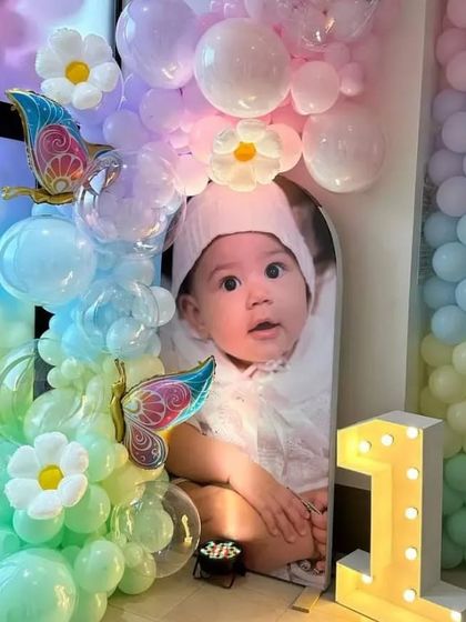 A precious and personalized setup for a first birthday. The decor features a large photo of the birthday baby, framed by a pastel rainbow balloon arch with butterfly and flower details, and a light-up number '1'.