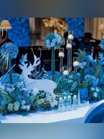A winter-themed table centerpiece. A white deer cutout is surrounded by blue hydrangeas, frosted foliage, and tall candle holders, perfectly matching the event's theme.