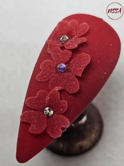 A beautiful example of 3D velvet-flocked flowers on a rich matte red base. The texture of the flowers contrasts with the smooth nail, and the rhinestone centers add a touch of sparkle.