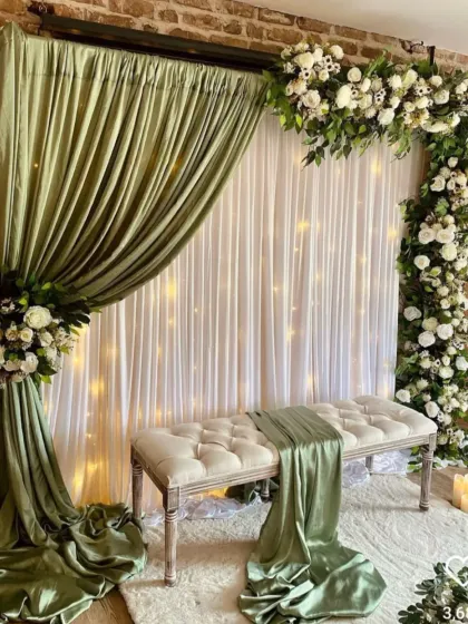 A very elegant and unique backdrop idea for a reception or engagement. This setup uses sage green drapes, an asymmetrical white floral arrangement, and a tufted bench for a soft, romantic feel.