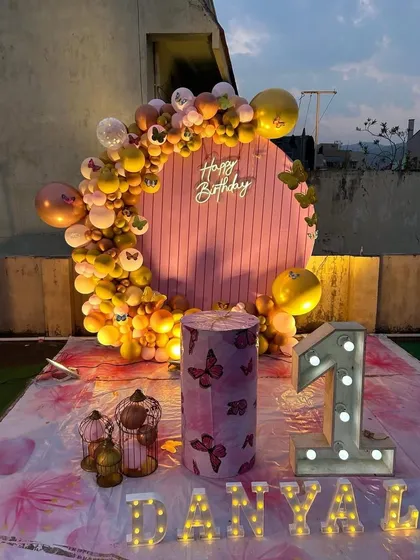 A first birthday celebration on a terrace with a pink circular backdrop. The backdrop is framed by a yellow and pink balloon garland, and the setup is completed with a themed plinth and light-up marquee numbers.