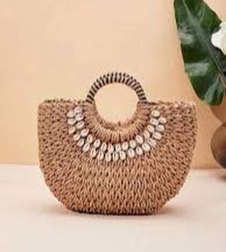 This semi-circular woven handbag is embellished with a delicate arc of cowrie shells, adding a bohemian and coastal touch. The design features a unique striped circular handle, making it a standout piece for any occasion.