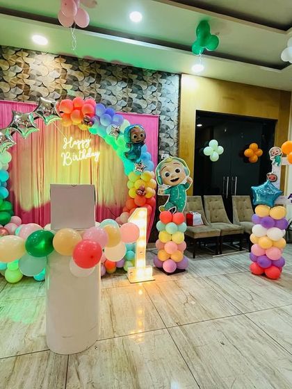 A large Cocomelon themed setup in a banquet hall. The decoration includes a colorful balloon arch, multiple character cutouts, and a light-up number '1'.