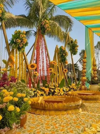 A grand outdoor Haldi setup with triangular backdrops made of marigold strings and sunflowers. The decor includes traditional lotus seats and a canopy of colorful drapes.