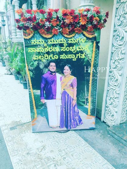 A personalized welcome board for a naming ceremony, featuring a photo of the parents. The sign is topped with a fresh flower arrangement.