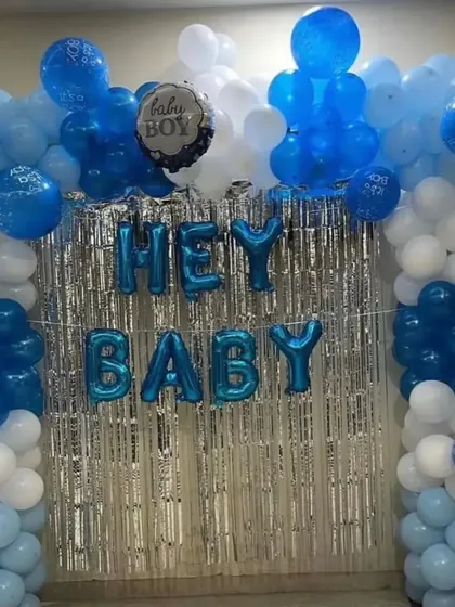 A classic 'Hey Baby' welcome decoration for a baby boy. The setup features a silver fringe backdrop framed by a blue and white balloon arch, with blue foil letters in the center.