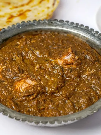 A close-up of a rich, dark Mughlai curry in a traditional serving bowl, paired with a paratha. The clean composition focuses on the texture and color of the gravy.