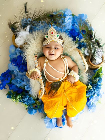 An adorable baby Krishna resting in a basket, surrounded by a sea of blue flowers and peacock feathers. This top-down shot is a creative and beautiful perspective.