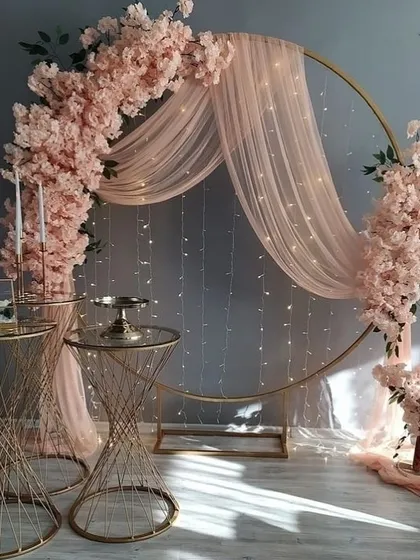 An inspiration for a romantic and elegant event setup, featuring a gold hoop arch with pink cherry blossoms, sheer drapes, and fairy lights.