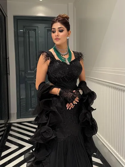 The structured updo adds height and elegance, balancing the voluminous ruffles of the gown and drawing attention to the emerald statement necklace.