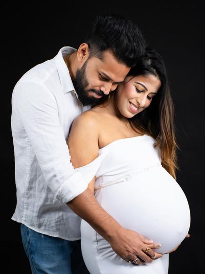 An intimate black and white portrait. The partner embraces the mom-to-be from behind, both looking down at the baby bump with love and wonder.