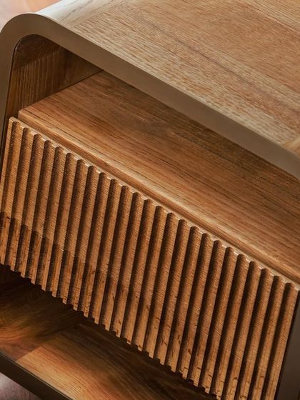 This bedside table features a fluted drawer front set within a gently curved metal and wood frame. The combination of sharp, vertical lines and soft, rounded corners creates a balanced and sophisticated piece.