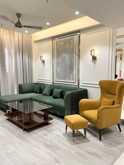 This living room setup combines a deep green L-shaped sofa with a contrasting mustard yellow wingback chair and ottoman. The classic wall moulding and modern sconce lighting add a layer of timeless elegance.