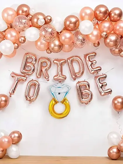A DIY-style "BRIDE TO BE" decoration against a simple white wall. This setup uses rose gold foil letter balloons, a diamond ring foil balloon, and a matching balloon garland, perfect for a simple and chic celebration at home.