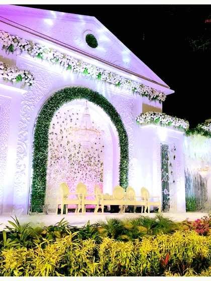 A closer look at the white palatial stage, where a green floral arch frames a waterfall and a backdrop of cascading flowers. The combination of textures creates a rich and dynamic visual experience.