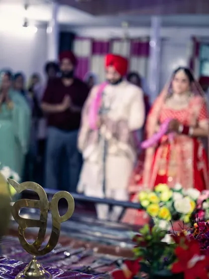 An artistic shot of the Anand Karaj ceremony, with the 'Ik Onkar' symbol in the foreground and the couple blurred in the background. This composition highlights the spiritual significance of the union.