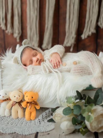 A full view of a charming setup featuring a baby in a knit outfit sleeping on a miniature bed, surrounded by tiny teddy bears and an alarm clock prop.