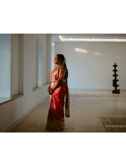 A bride in a beautiful red saree, standing in a minimalist, well-lit hall. The soft light from the window creates a peaceful and serene portrait.
