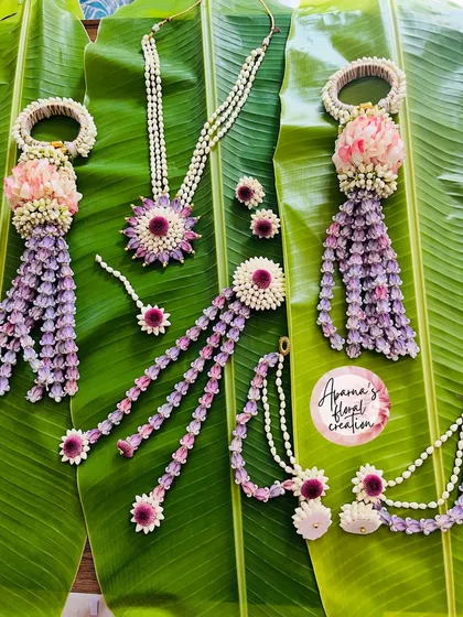 A vibrant floral jewellery set perfect for a Haldi or Mehendi ceremony, laid out on a banana leaf. This design combines purple and pink flowers with strings of white buds, and includes a necklace, maang tikka, earrings, and hath phool.