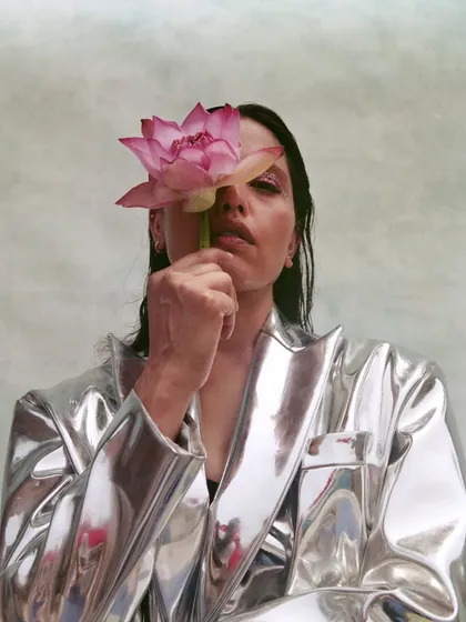 A conceptual portrait where a metallic silver jacket is paired with a natural element, a pink lotus. The styling creates a beautiful contrast between the hard, reflective surface of the jacket and the soft, organic texture of the flower.