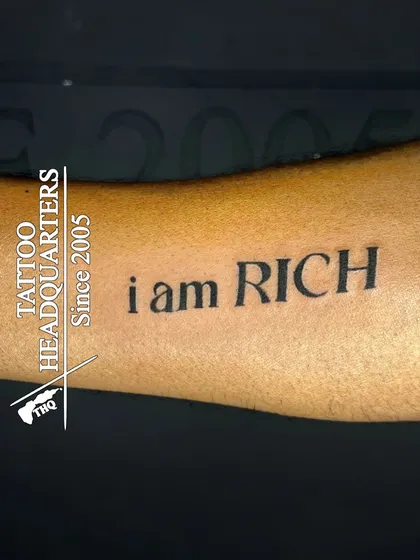 A bold statement tattoo that reads "i am RICH" in a classic serif font on the forearm.