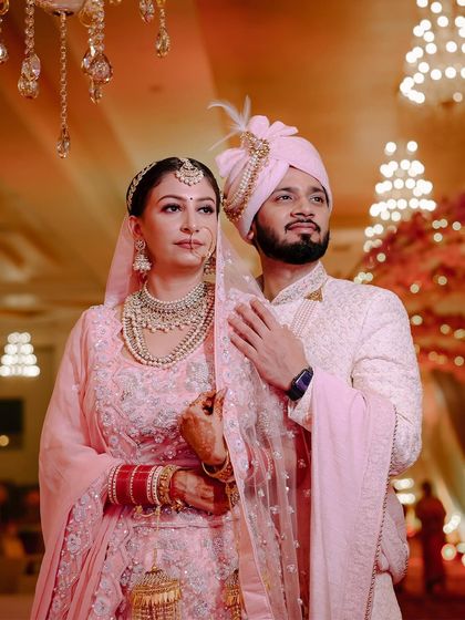 A classic portrait of a couple on their wedding day in Jammu. Their pastel pink outfits and the grand, warmly lit venue create an elegant and romantic image.