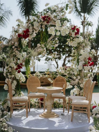 A close-up of an intimate mandap setup by the pool, featuring a deconstructed arch of white and red flowers. The organic, free-flowing floral design creates a romantic and natural frame for the ceremony, perfect for a modern garden wedding.