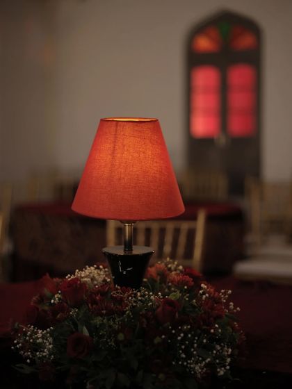 A moody detail shot from the Sangeet. A simple red table lamp on a floral centerpiece adds a warm, intimate glow to the red velvet-draped tables.