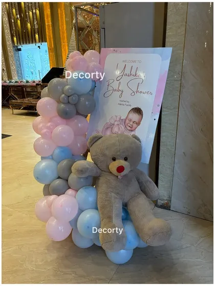 A cute welcome display for a baby shower, featuring a personalized sign, a teddy bear, and a pink and blue balloon arrangement.